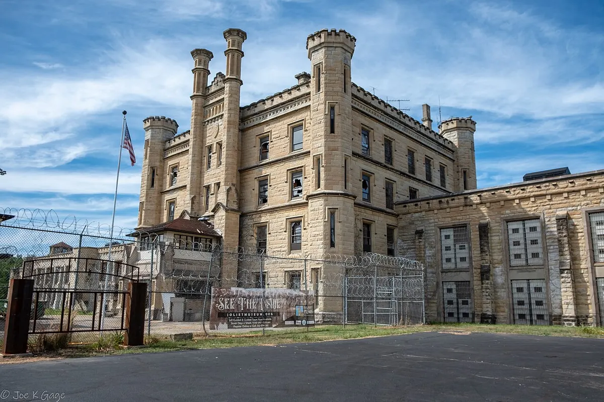 The turreted limestone administration building of Old Joliet Prison behind razor-wire fencing, with a Joliet Area Historical Museum tour banner