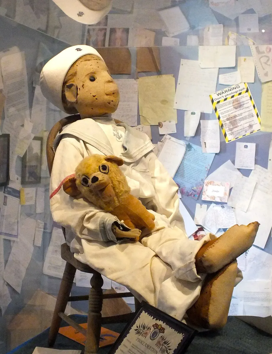 Robert the Doll in his white sailor suit, seated in his display case holding a toy lion